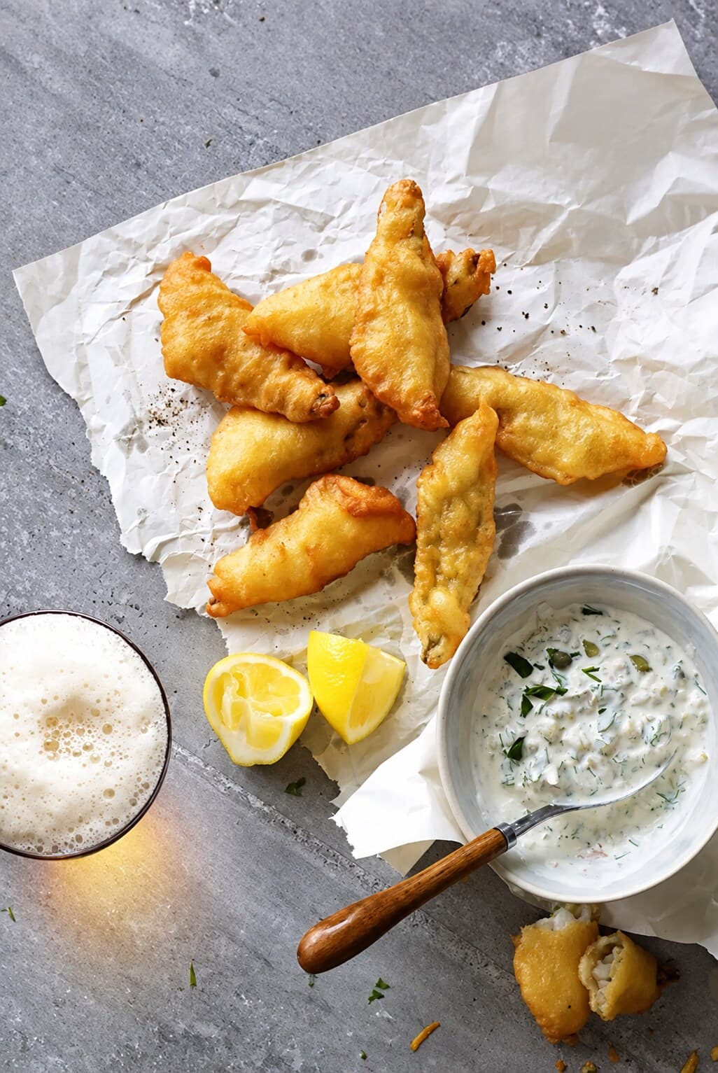 Frittierte Fisch-Knusperli auf Backpapier, dazu Zitronenschnitze, eine Sauce tartare und ein Glas Bier