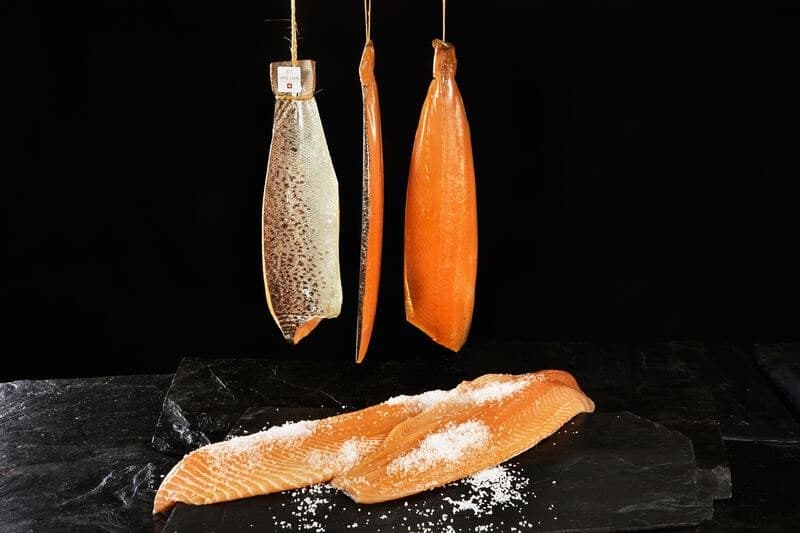 Drei hängende Lachsseiten mit dem Etikett von Swiss Lachs, davor eine gesalzene Lachsseite auf Schiefer